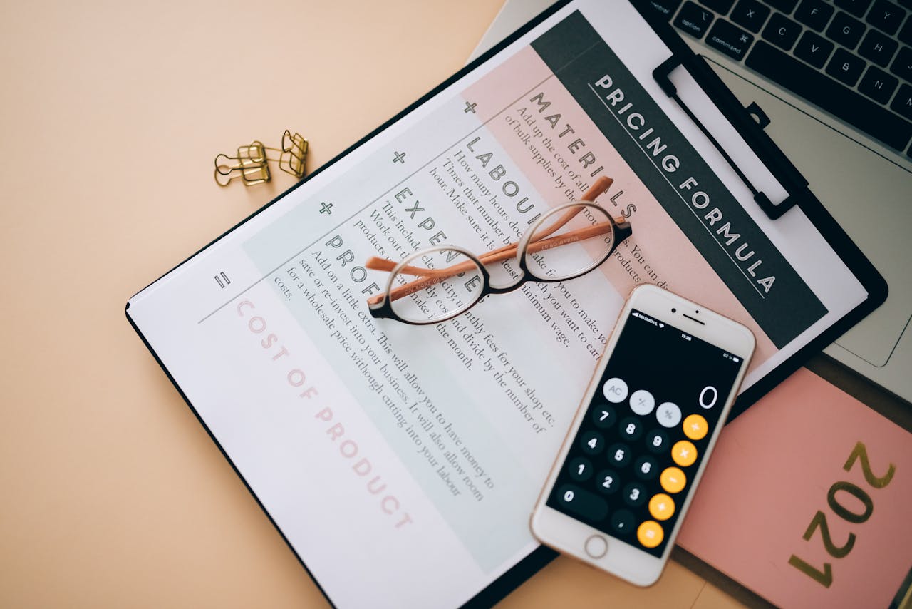 Overhead view of business tools including a phone calculator, pricing formula document, and eyeglasses on a desk.